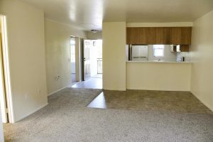 Carpeted living room with neutral color walls overlooking kitchen, front door and tiled dining area